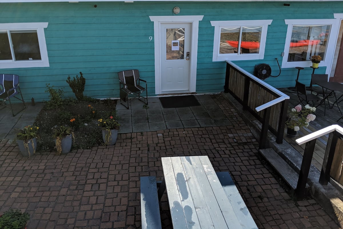 A turquoise building with a brick patio, a white door, and a small garden with potted plants.