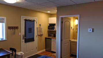 A compact bedroom with a bed, a desk, a chair, a microwave, and a small kitchenette.
