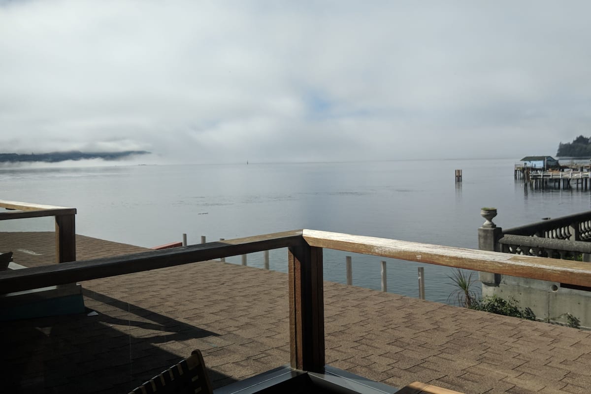 A waterfront view from a wooden deck with a railing, overlooking calm water and a distant shoreline.