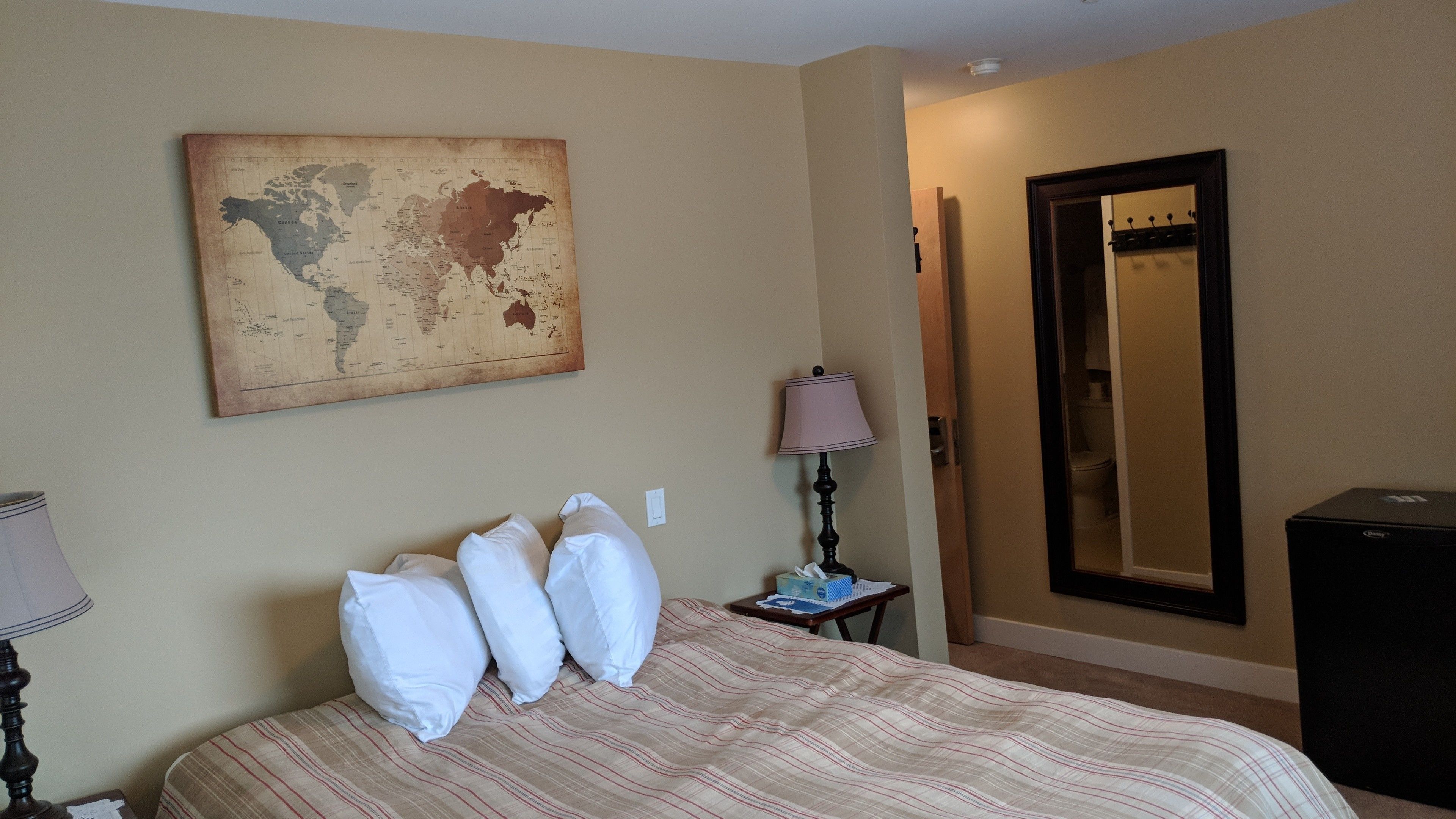 A hotel room with a bed, pillows, a map on the wall, a lamp, a mirror, and a dresser.