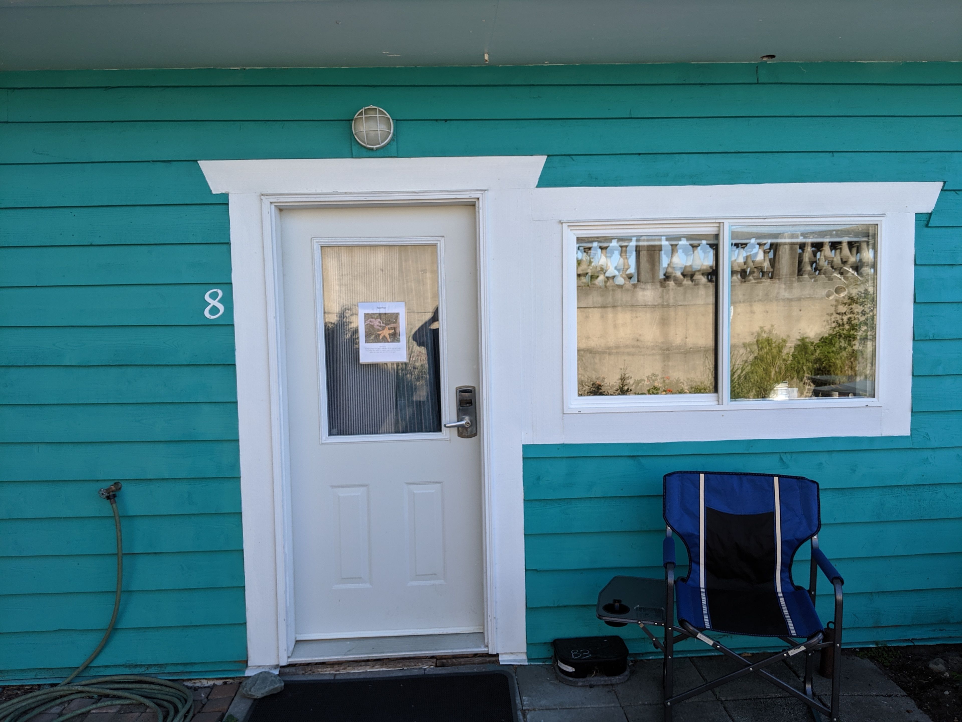 A teal house with a white door, a window with a view of a concrete wall, and a blue folding chair outside.