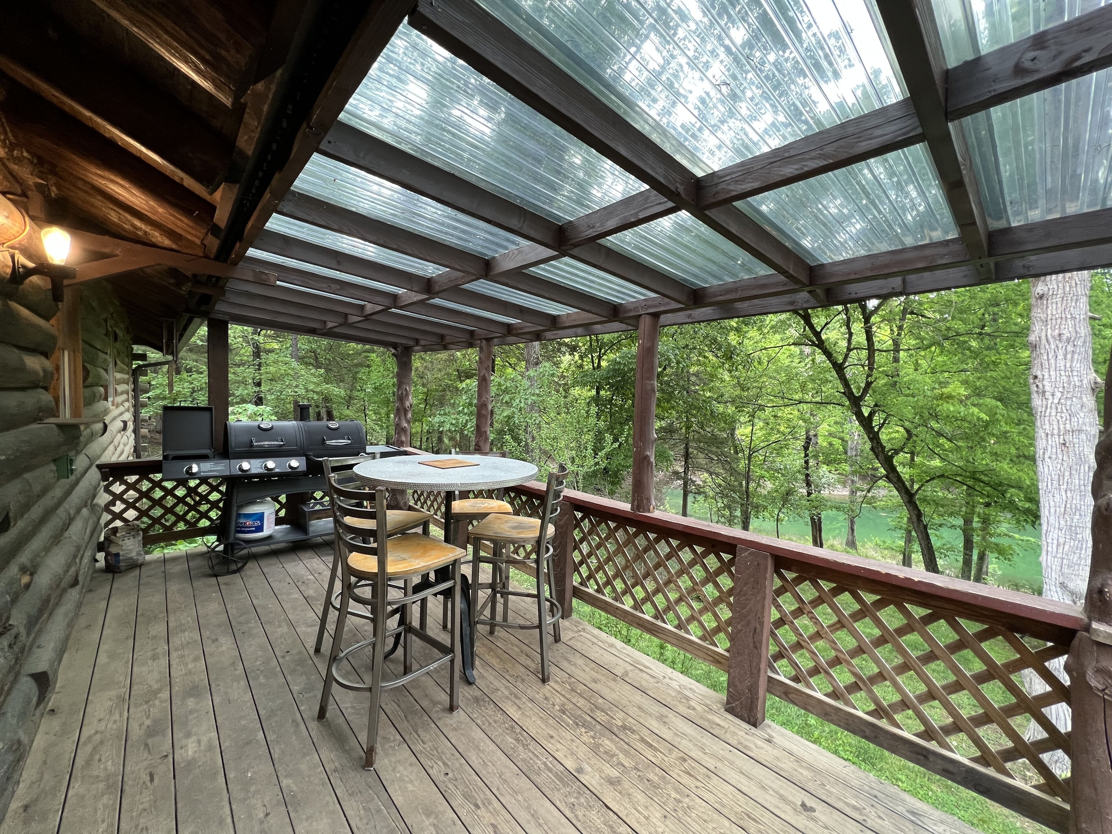 Eine überdachte Terrasse im Freien mit Grill, Tisch und Stühlen, umgeben von einem Holzzaun und Bäumen.