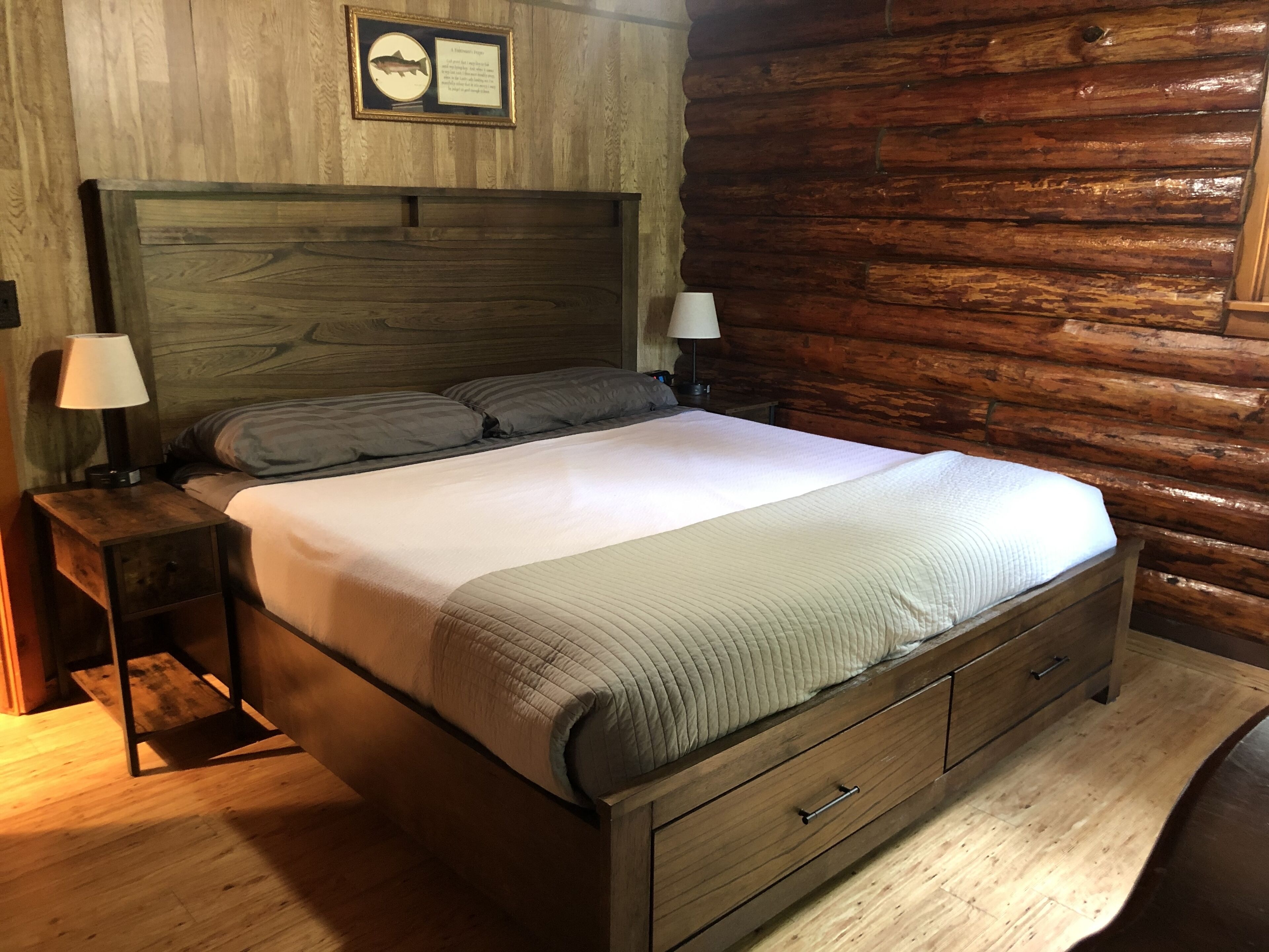 A wooden log cabin-style bedroom with a large bed, two bedside tables with lamps, and a framed fish picture on the wall.