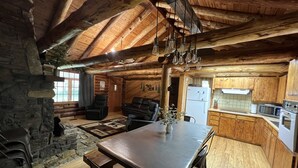 A rustic cabin interior with a stone wall, wooden ceiling, a dining area, a kitchen with wooden cabinets, and a fireplace. - Cozy Lake Front Family Friendly Cabin, Bunk House, Kayaks and Canoe! (Rogers)