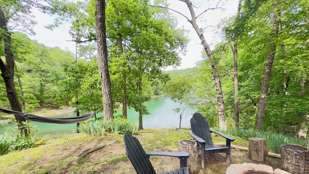 A serene outdoor seating area with Adirondack chairs and a fire pit, overlooking a lake surrounded by lush greenery. - Cozy Lake Front Family Friendly Cabin, Bunk House, Kayaks and Canoe! (Rogers)