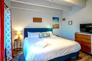 Ein ordentlich bezogenes Bett mit weißen Leinen, einem blauen Kopfteil und dekorativen Kissen. Ein Fernseher ist an einem Holzschrank rechts montiert.