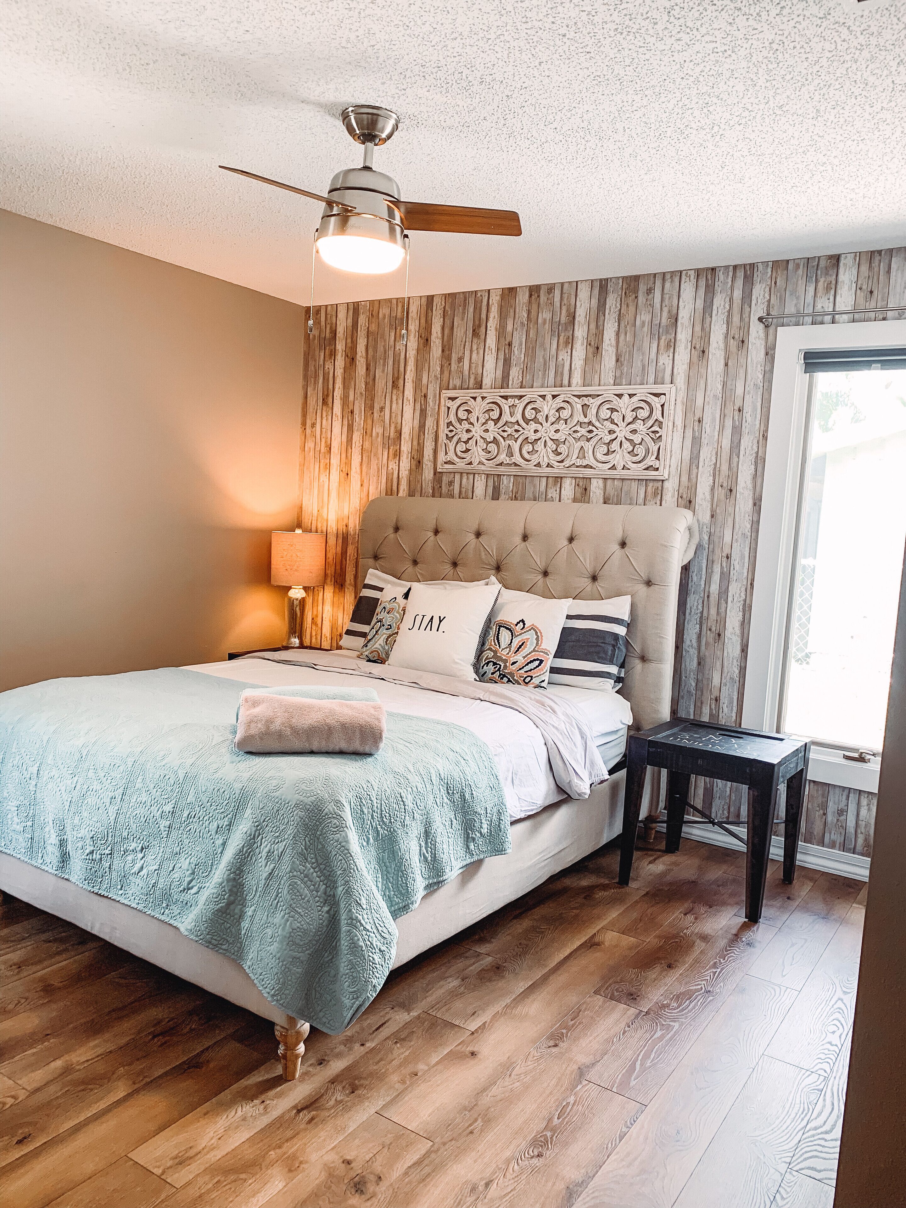 A bedroom with a tufted headboard, a bed with patterned bedding, a nightstand with a lamp, and a wooden floor.