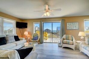 A living room with a sofa, armchairs, a glass coffee table, and a ceiling fan. There is a large window with blinds, a flat-screen TV mounted on the wall, and a view of the beach through the glass door. - Experience ocean views from this spacious 4-bedroom second row home in Surf City! (Surf City)