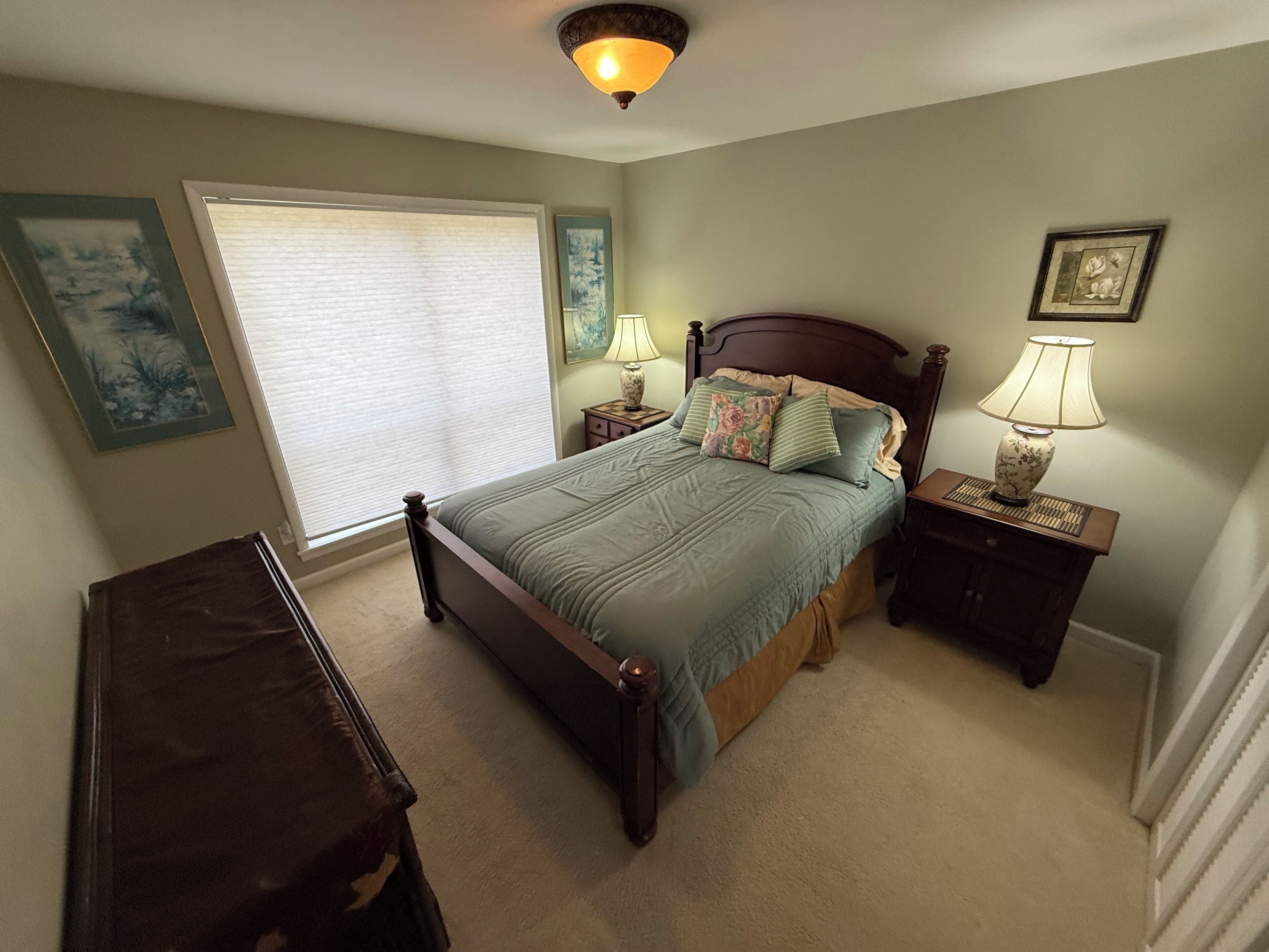 A bedroom with a bed, nightstands, a dresser, a window with blinds, and wall art.