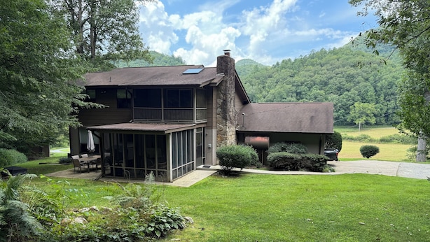 A two-story house with a covered patio, stone exterior, and a view of a lush green landscape. - Large private home on wooded acreage (Robbinsville)