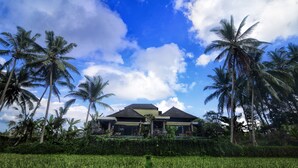 A two-story house with a balcony, surrounded by a lush green field and palm trees under a partly cloudy sky. - Villa Kirani Ubud (Ubud)