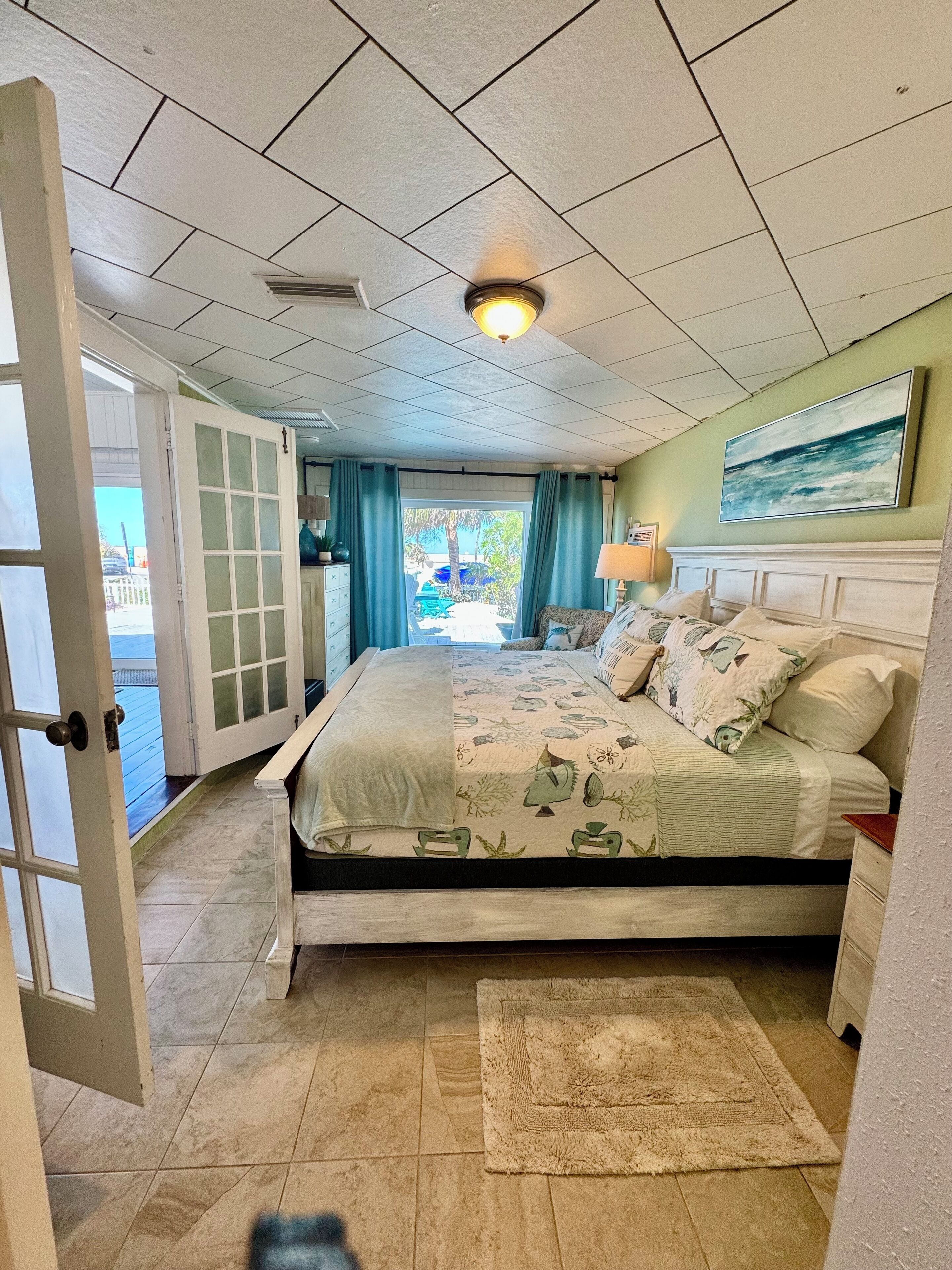 Un dormitorio con cama, mesita de noche, lámpara, un cuadro en la pared y vistas al exterior.