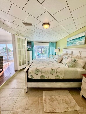 A bedroom with a bed, a nightstand, a window with curtains, and a view of the outdoors.