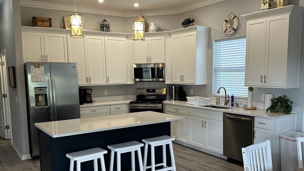A modern kitchen with white cabinets, stainless steel appliances, a central island with bar stools, and a dining area with a wooden table and white chairs. - Sea Shell Haven 
Family Home, 300 yards from the beach, 2 private docks, Grill! (Placida)