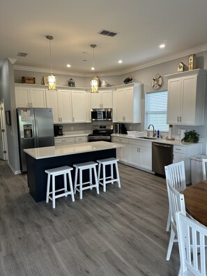 Een moderne keuken met witte kastjes, roestvrijstalen apparatuur, een centraal eiland met barkrukken en een eetgedeelte met een houten tafel en witte stoelen.