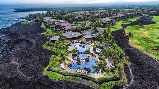 An aerial view of a resort with multiple buildings, a central pool area, and a surrounding landscape with a coastline. - Ocean View Escape – Poolside Calm & Weekly Savings (Waikoloa)