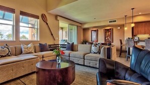 A modern living room with a sectional sofa, a round coffee table, and a dining area with a kitchen in the background. - Ocean View Escape – Poolside Calm & Weekly Savings (Waikoloa)