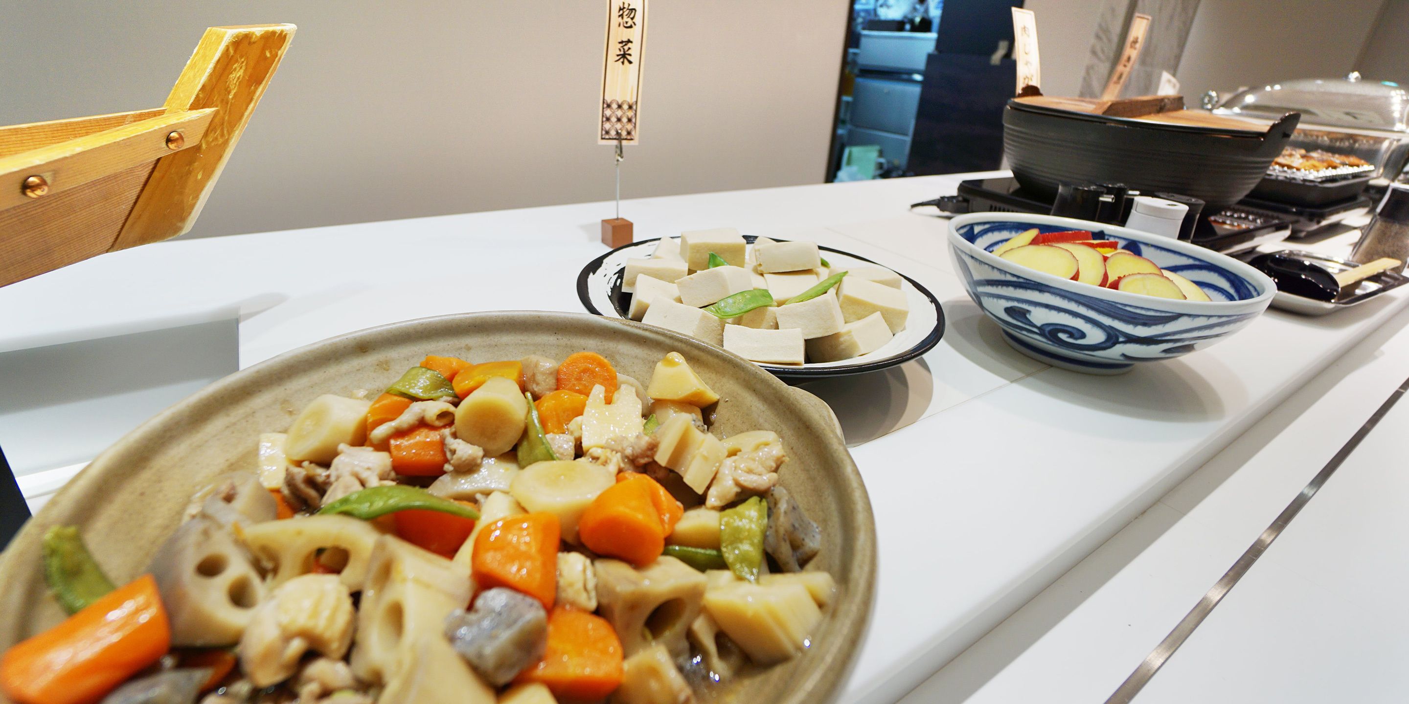 A buffet table with various dishes including tofu, vegetables, and a bowl of fruit.