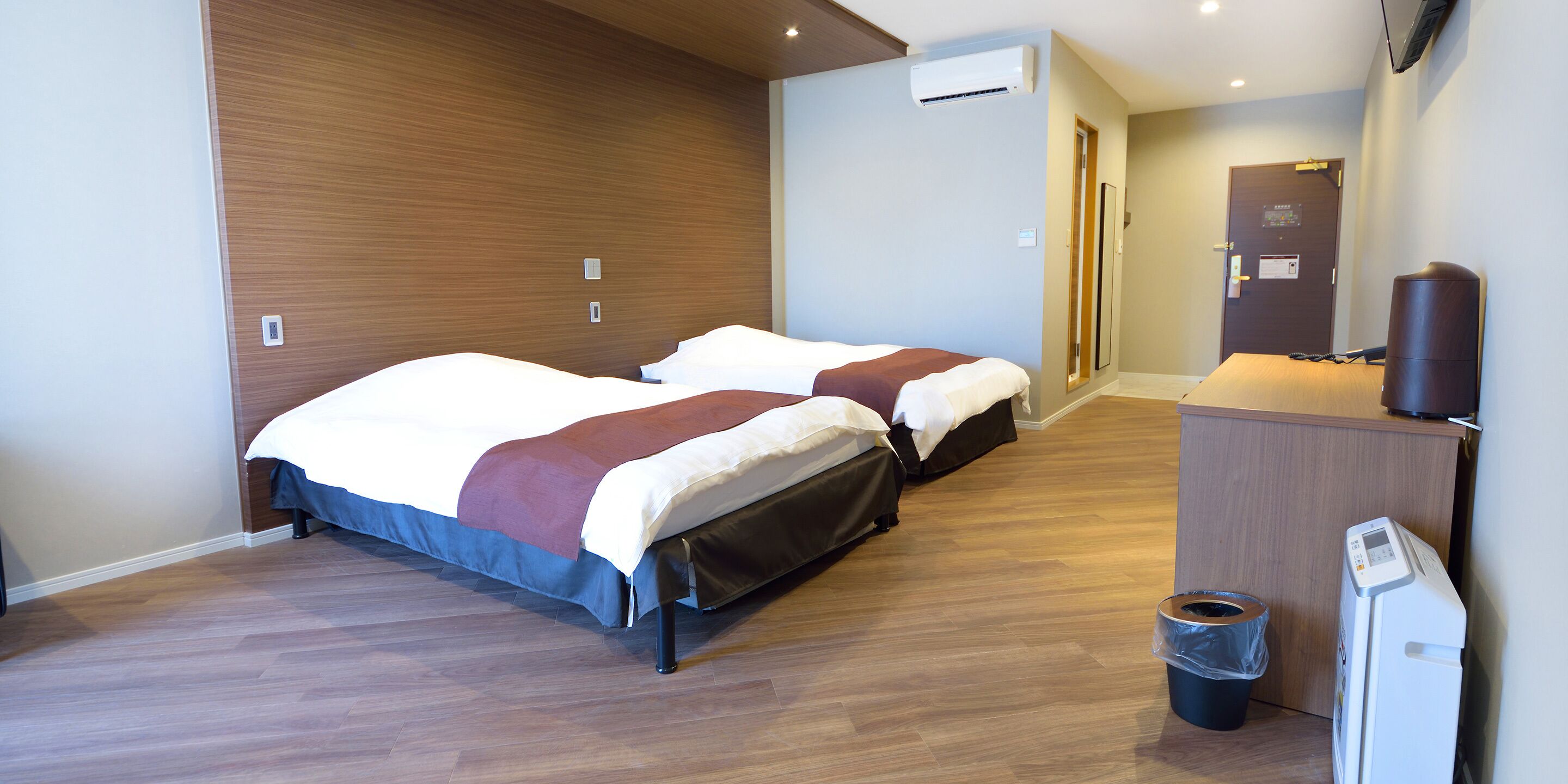 Une chambre d’hôtel avec deux lits, un plancher en bois, un appareil de climatisation, un bureau avec une poubelle et une porte donnant sur une autre chambre.
