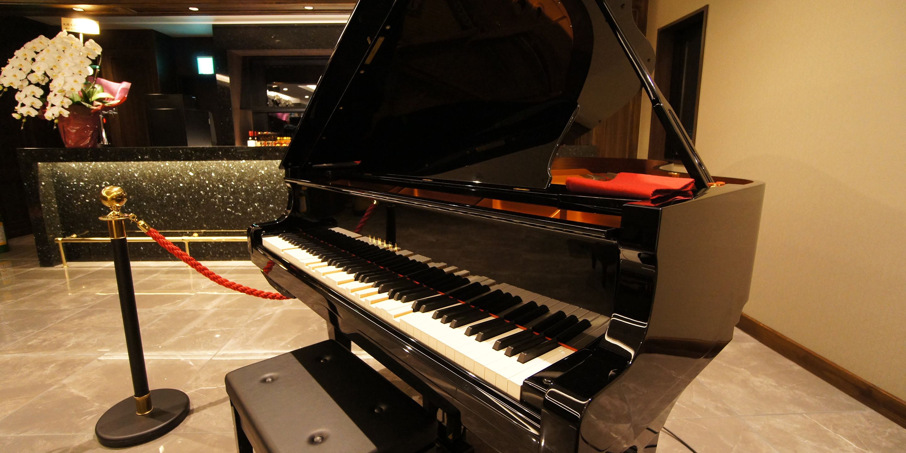A grand piano in a lobby with a marble floor, a red rope barrier, and a vase of white flowers.