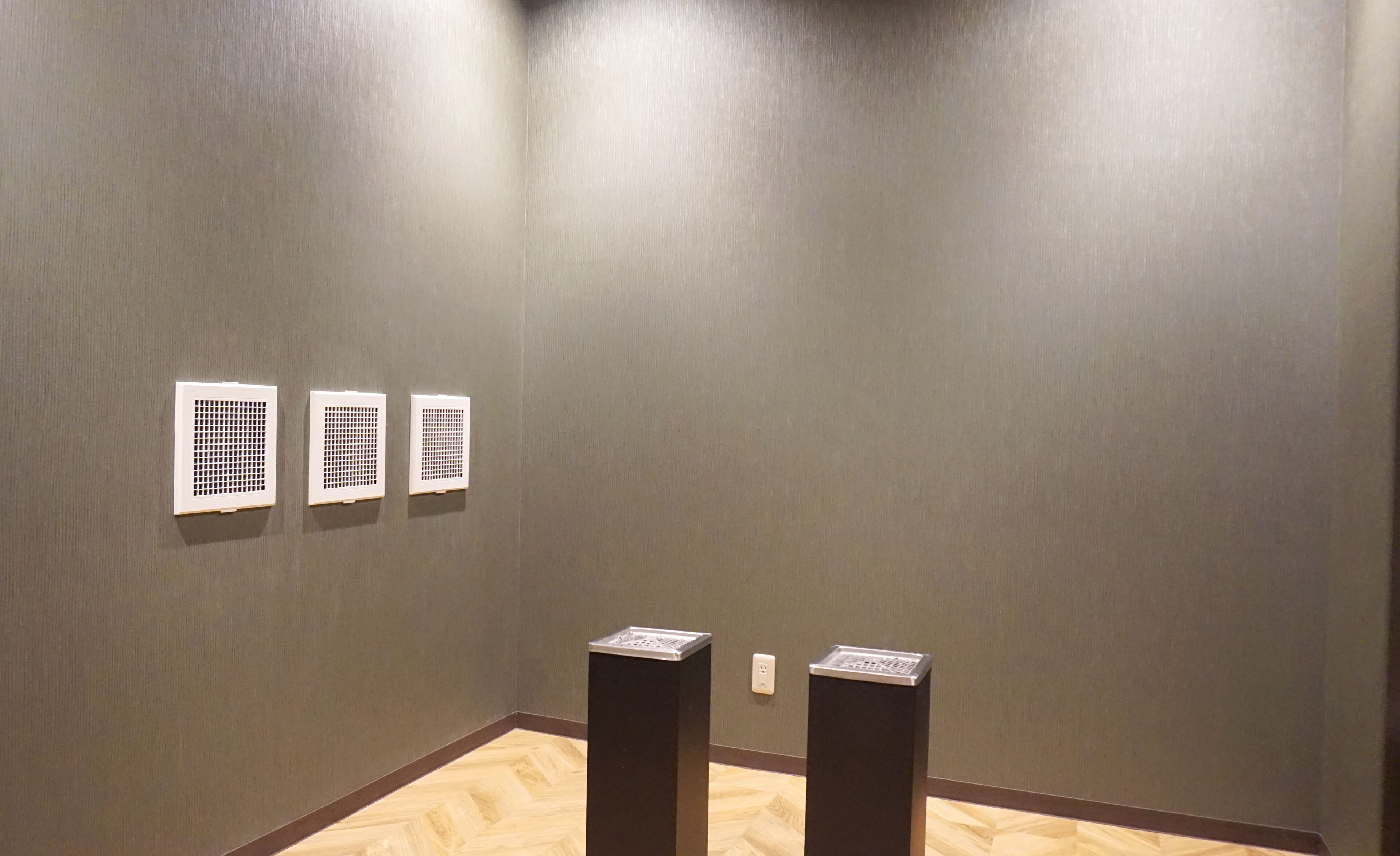 A hallway with a grey wall, three white vents, and two black pillars.