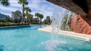 Une piscine agrémentée d’une cascade, entourée de palmiers et d’un mur de pierre.