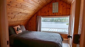 A cozy bedroom with a wooden ceiling, a large bed with a gray bedspread, and a window overlooking a body of water. - Cottage - Lakeside w/private Hottub near Big White (Naramata)