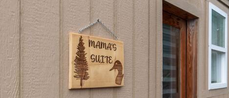 A wooden sign with a pine tree and a snake, labeled "MAMA'S SUITE," hanging on a beige wall.