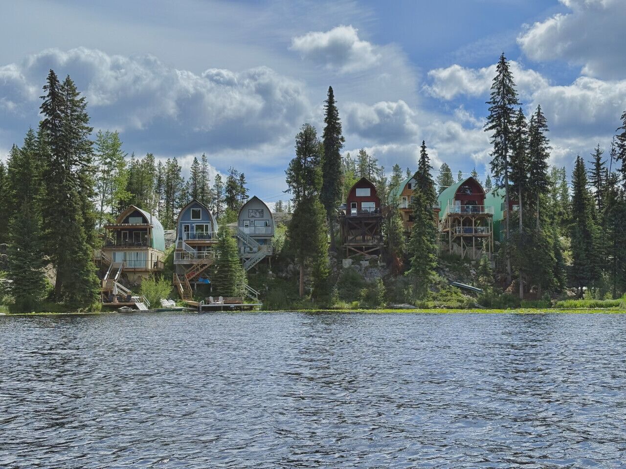 A lakeside cabin complex with multiple buildings, including a large house with a wrap-around porch and a smaller cabin with a steep staircase.