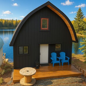 Une cabane noire avec une terrasse en bois, deux chaises Adirondack bleues et une table ronde en bois.