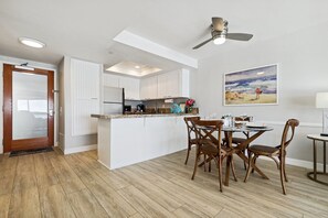 A modern kitchen with white cabinets, a central island, and a dining area with wooden chairs. - 1 Bedroom Condo on the Sand! (Carlsbad)