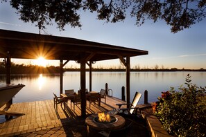 A wooden deck with a fire pit, outdoor seating, and a view of a calm lake at sunset.