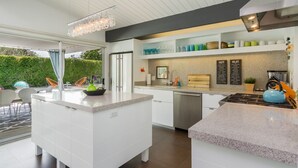 Cocina moderna con armarios blancos, encimera de granito y estufa integrada. Hay una zona de comedor con mesa y sillas, y un ventanal con vistas al exterior.