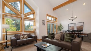 A spacious living room with large windows, a brown leather sofa, a coffee table, and a dining area with a wooden table and chairs. - Timber Lake 341 Private outdoor hot tub, views, garage parking (Winter Park)