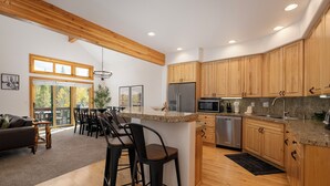 A modern kitchen with wooden cabinets, granite countertops, and stainless steel appliances. There is a dining area with a large window and a view of trees. - Timber Lake 341 Private outdoor hot tub, views, garage parking (Winter Park)