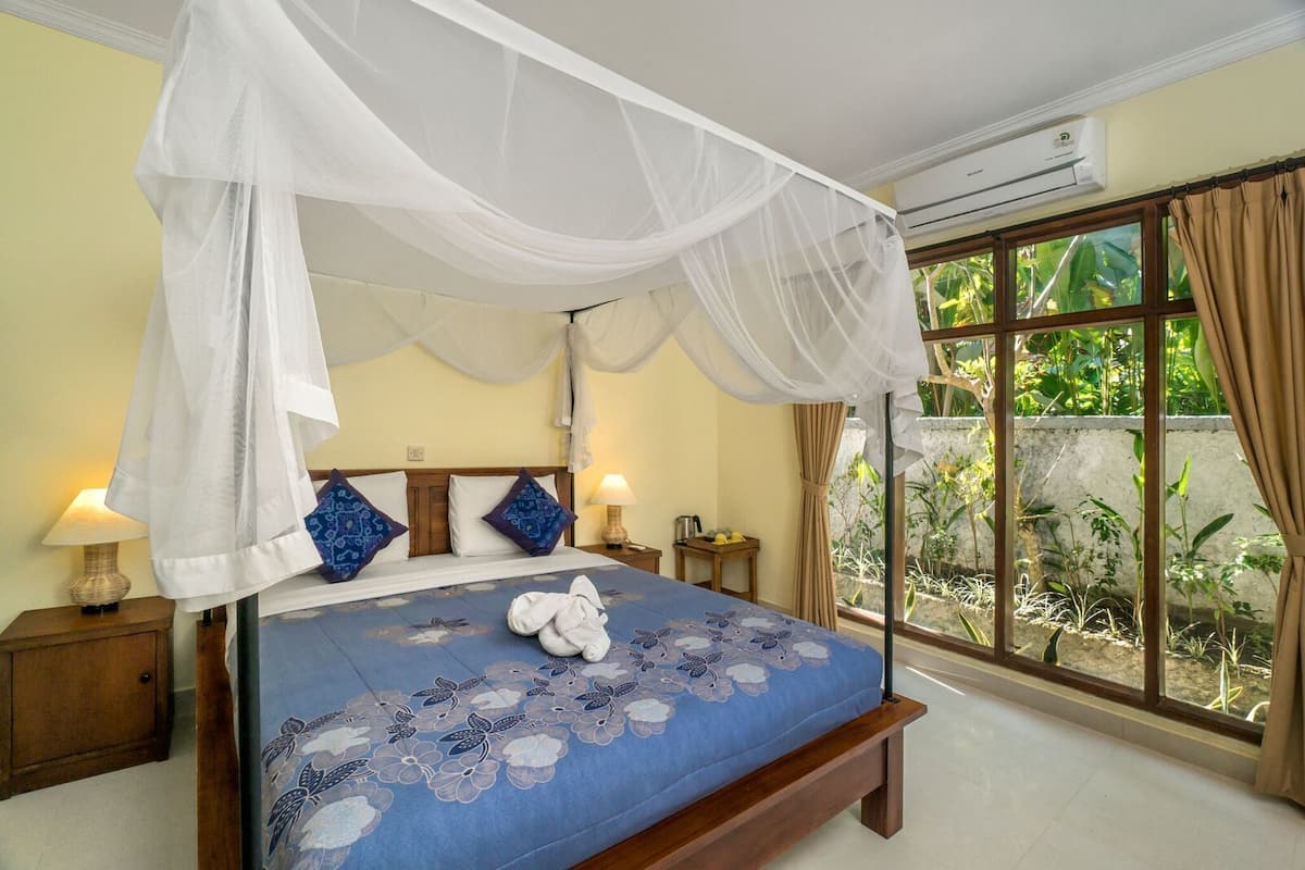 A bedroom with a four-poster bed, a canopy, a large window with curtains, and a view of greenery outside.