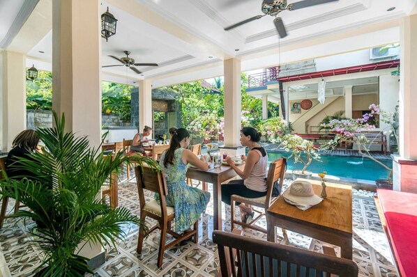 Restaurante com área externa e mesas, cadeiras de madeira, vasos de plantas e ventilador de teto, com vista para uma piscina.