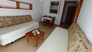 A living room with a sofa, a coffee table with cups and a coffee pot, and a dining area with a table and chairs. - APCOSTAS Lotus / One Bedroom (Blanes)