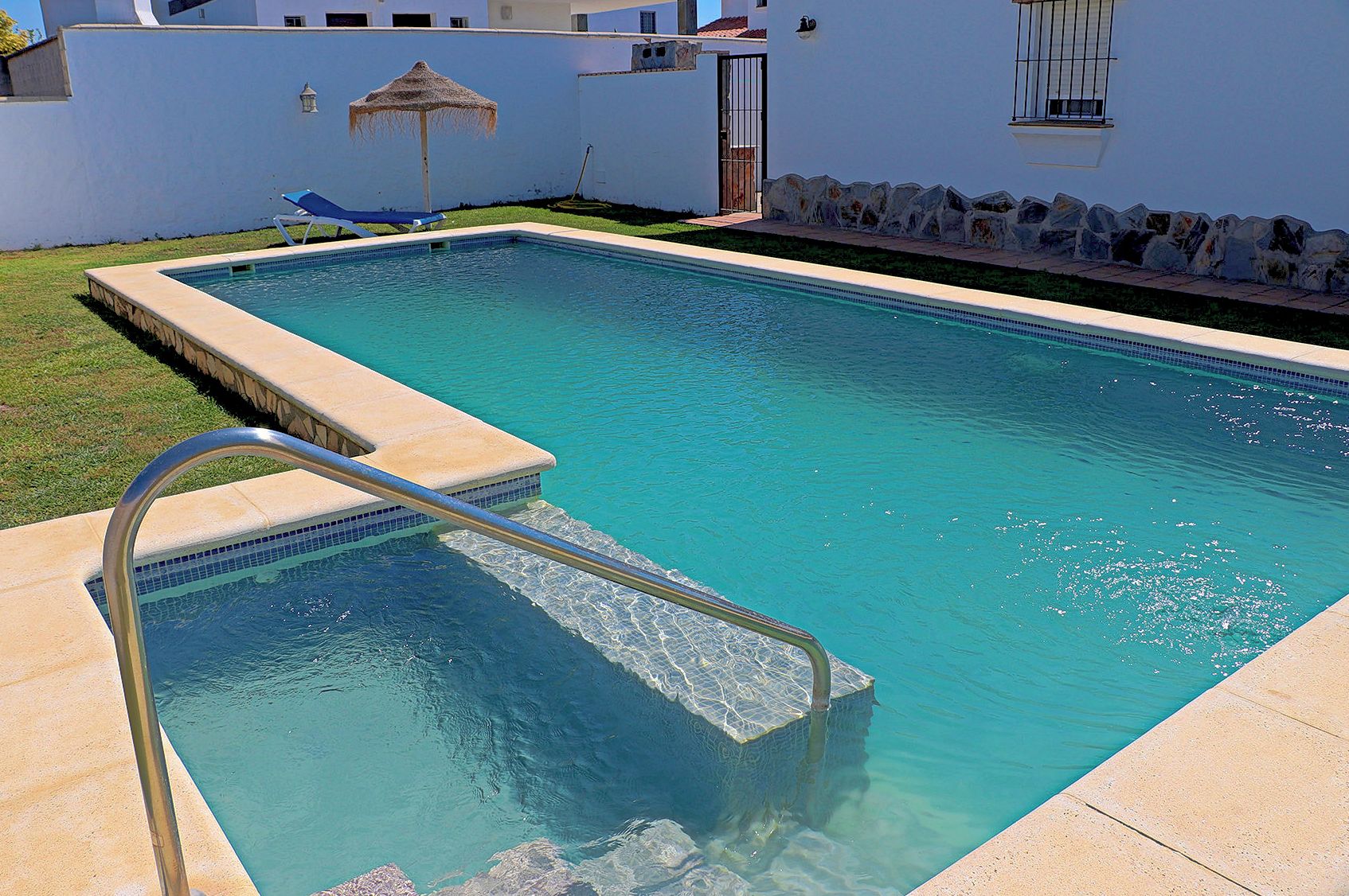 Ein rechteckiger Swimmingpool mit einer Metallleiter, einem Reedschirm und einer Steinmauer mit einer Steinumrandung.