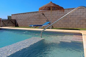 Ein Swimmingpool mit einer Metallleiter, einer blauen Liege und einer Mauer mit einer strohgedeckten Struktur.
