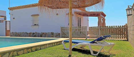Una piscina con sombrilla de paja, zona vallada y edificio con pared de piedra y ventanas.