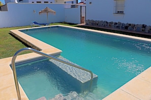 Una piscina rettangolare con una scala in metallo, un ombrellone di paglia e un muro di pietra con borda in pietra.