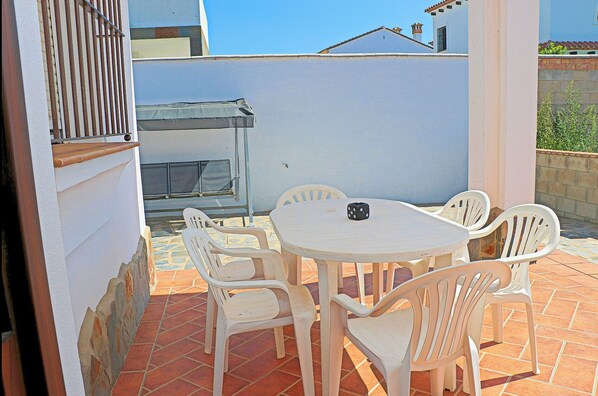 Eine Terrasse mit einem runden Tisch und weißen Plastikstühlen, ein kleines schwarzes Objekt auf dem Tisch und im Hintergrund ein Gebäude.