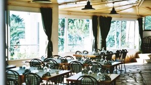 Een restaurant met meerdere tafels en stoelen, grote ramen met gordijnen en een vloer van stenen tegels.