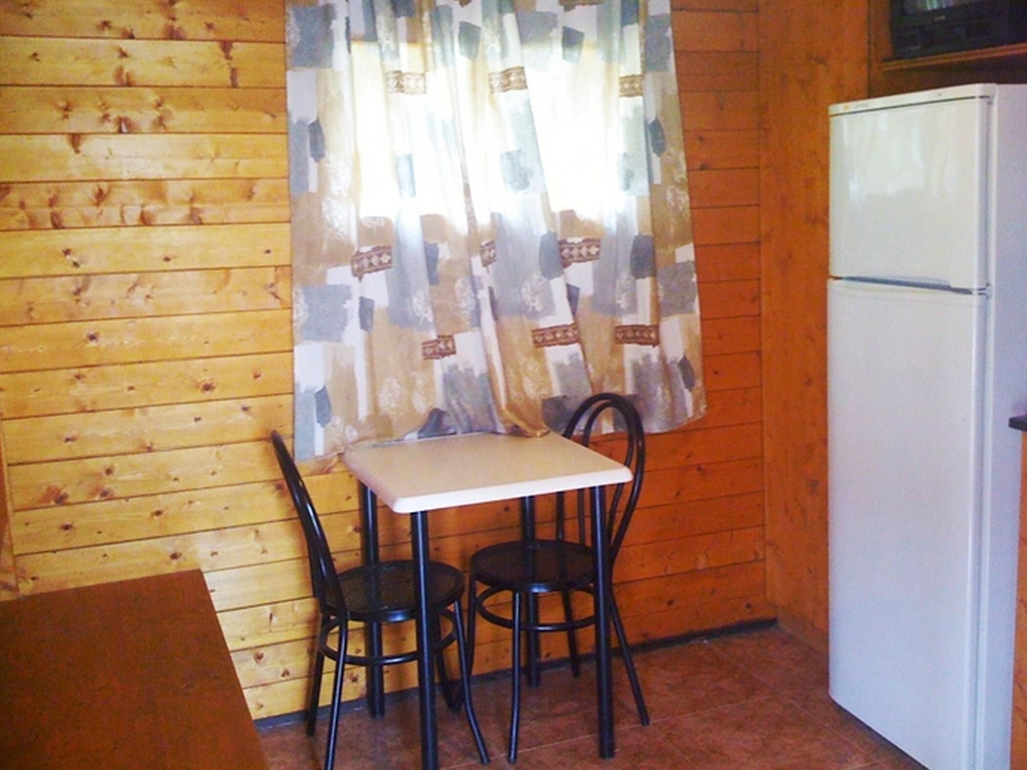 Una cocina pequeña con pared de madera, un refrigerador blanco, una mesa con dos sillas y una cortina estampada.