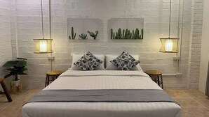 A bedroom with a white brick wall, a bed with white bedding and floral pillows, two wall-mounted cactus displays, and two hanging lamps. - Yindee Travellers Lodge (Nan)
