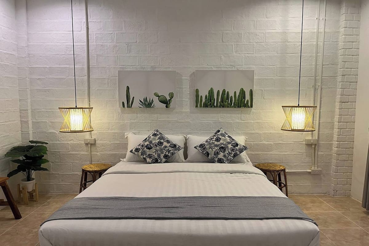 Ein Schlafzimmer mit weißer Ziegelwand, einem Bett mit weißer Bettwäsche und floralen Kissen, zwei Wanddekorationen mit Kakteen und zwei Pendelleuchten.