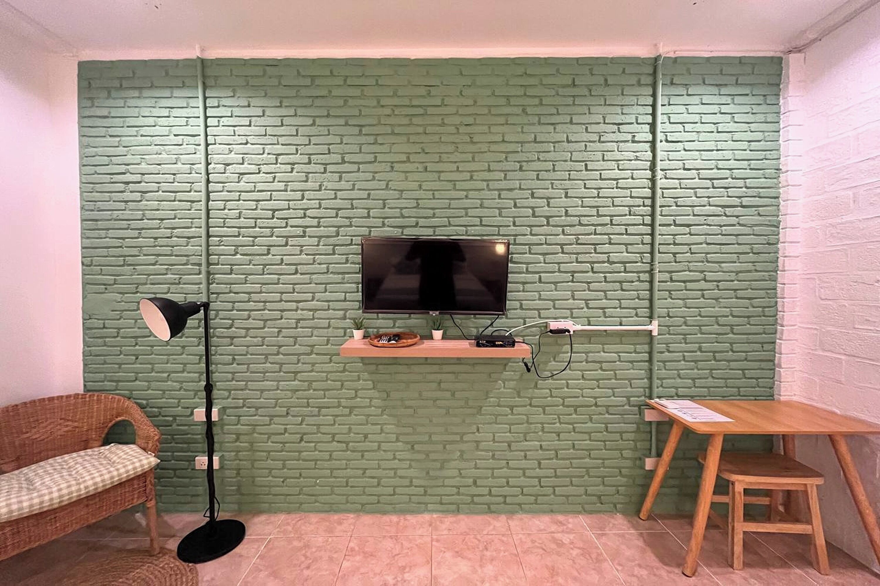 A room with a brick-patterned wall, a mounted TV, a floor lamp, a small table, and a wicker sofa.