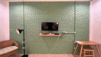 A room with a brick-patterned wall, a mounted TV, a floor lamp, a small table, and a wicker sofa.