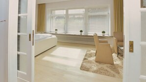 Et moderne soveværelse med en stor seng, et skrivebord og en stol. Der er vinduer med persienner og en radiator under vinduerne.